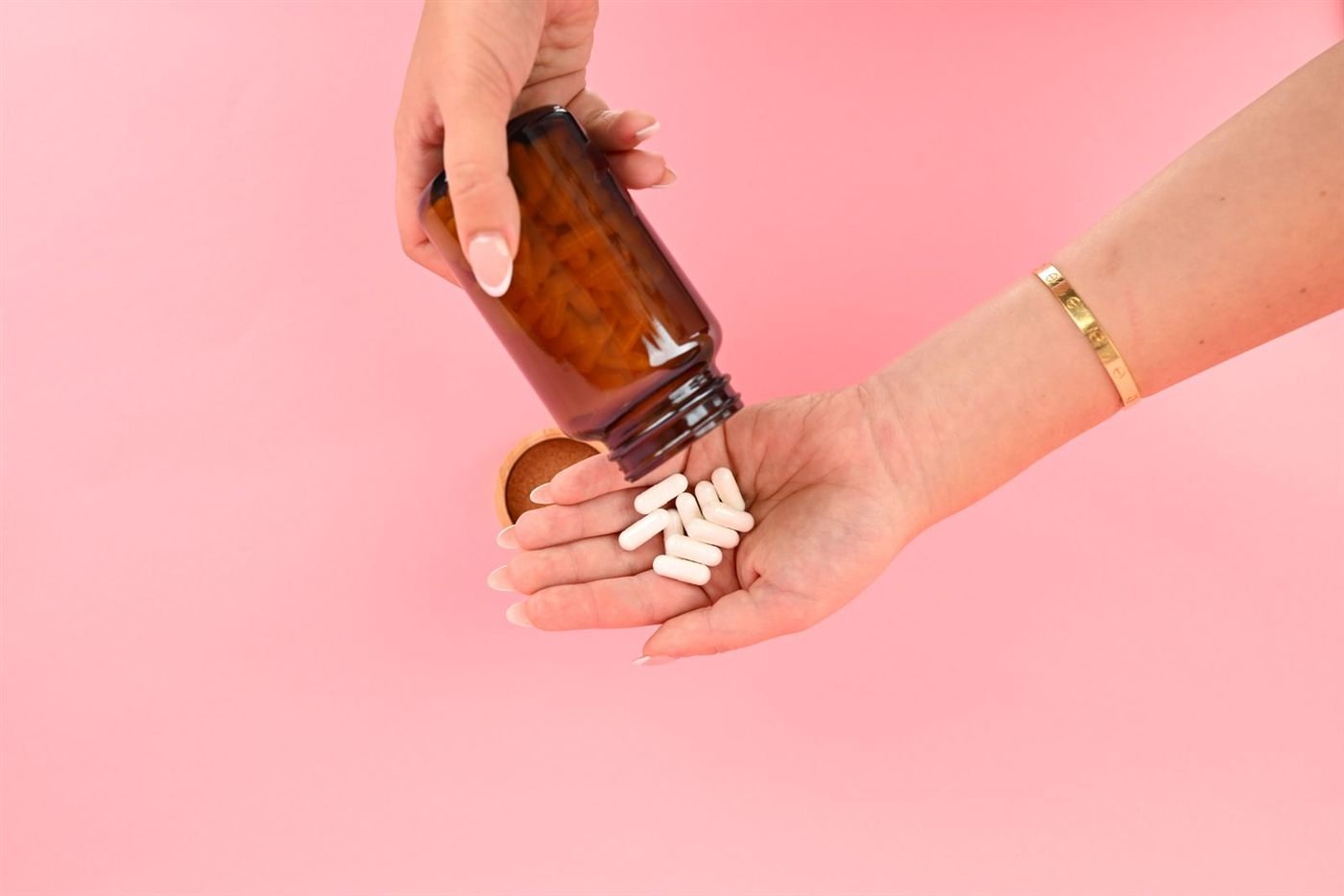Mão derramando cápsulas de suplemento de um frasco sobre fundo rosa