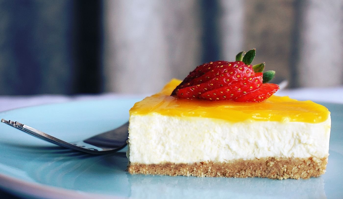 Fatia de cheesecake fit coberta com morango fresco e calda, servida em prato azul com garfo ao lado