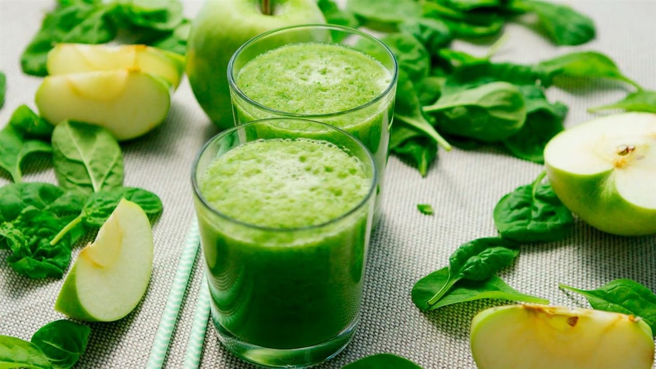 Dois copos de smoothie verde de maçã e espinafre cercados de folhas e fatias de maçã